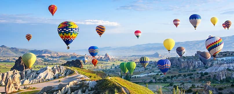 Viele-bunte-Heissluftballons-schweben-ueber-einer-felsigen-und-gruenen-Landschaft-mit-blauem-Himmel