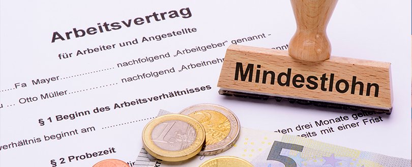 Mindestlohngesetze - TEGA GmbH Bargeld-und-Stempel-mit-der-Aufschrift-Mindestlohn-liegen-auf-einem-Arbeitsvertrag-Dokument